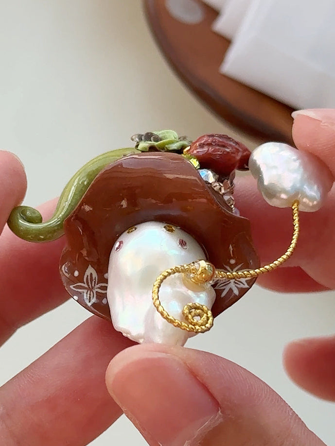 [PIECE10] Bakery Sprite baroque pearl necklace with taupe bagel-inspired hat, sculpted bagel detail, floral accents, gold wand, and natural baroque pearl
